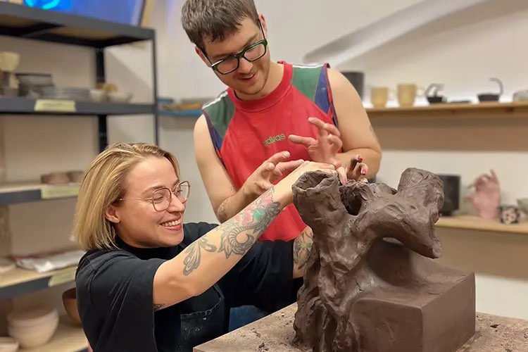 Two people laughing as they build a sculptural piece in dark clay